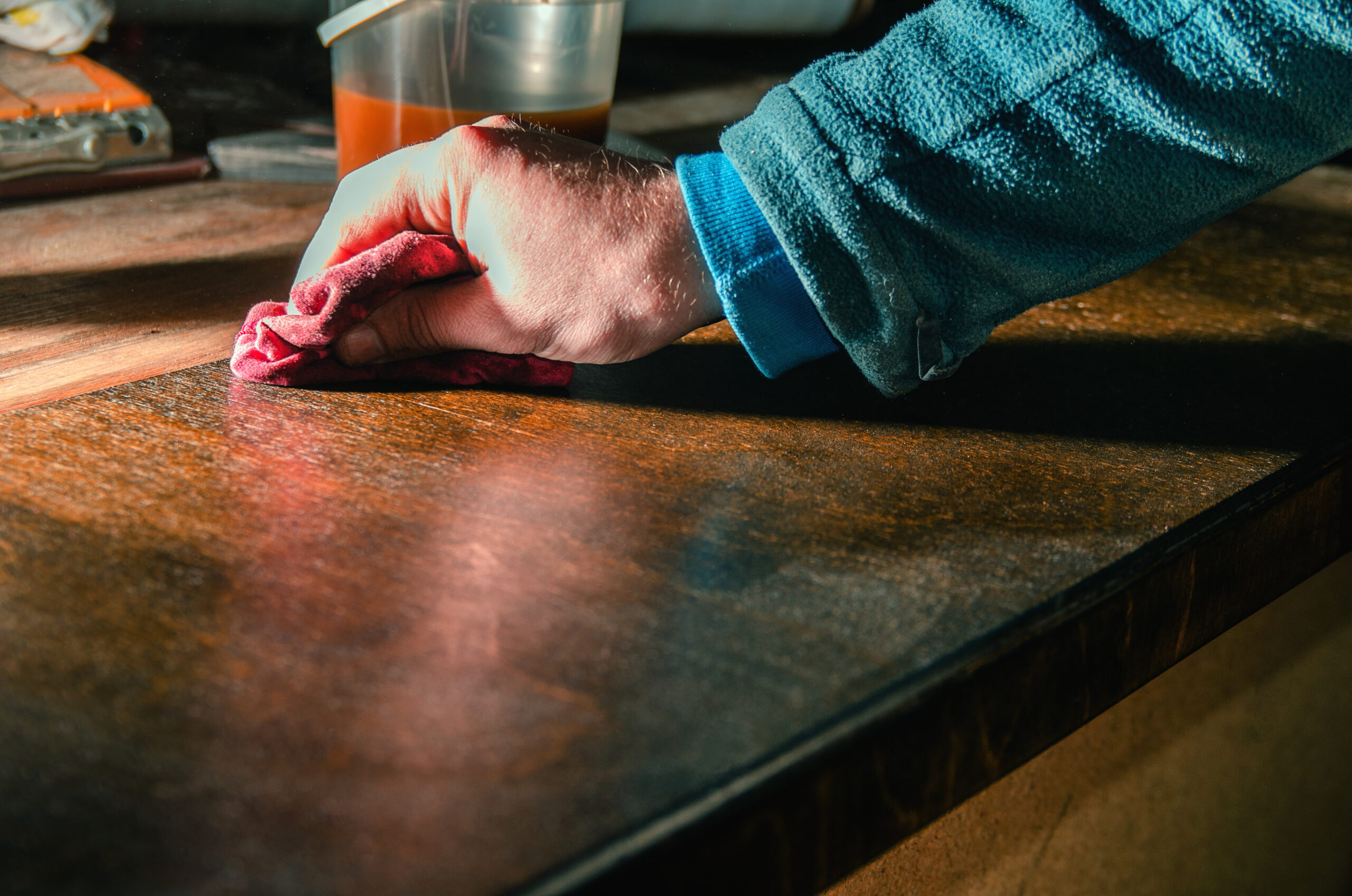 A hand is applying coating onto a piece of wood with a red rag. The parts of the table he's rubbed are noticeably sleeker.