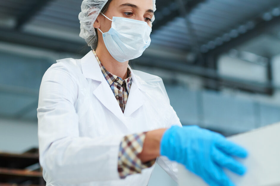 An employee wearing a mask and blue gloves is handling a white cardboard box. The box has a clear matte sheen.