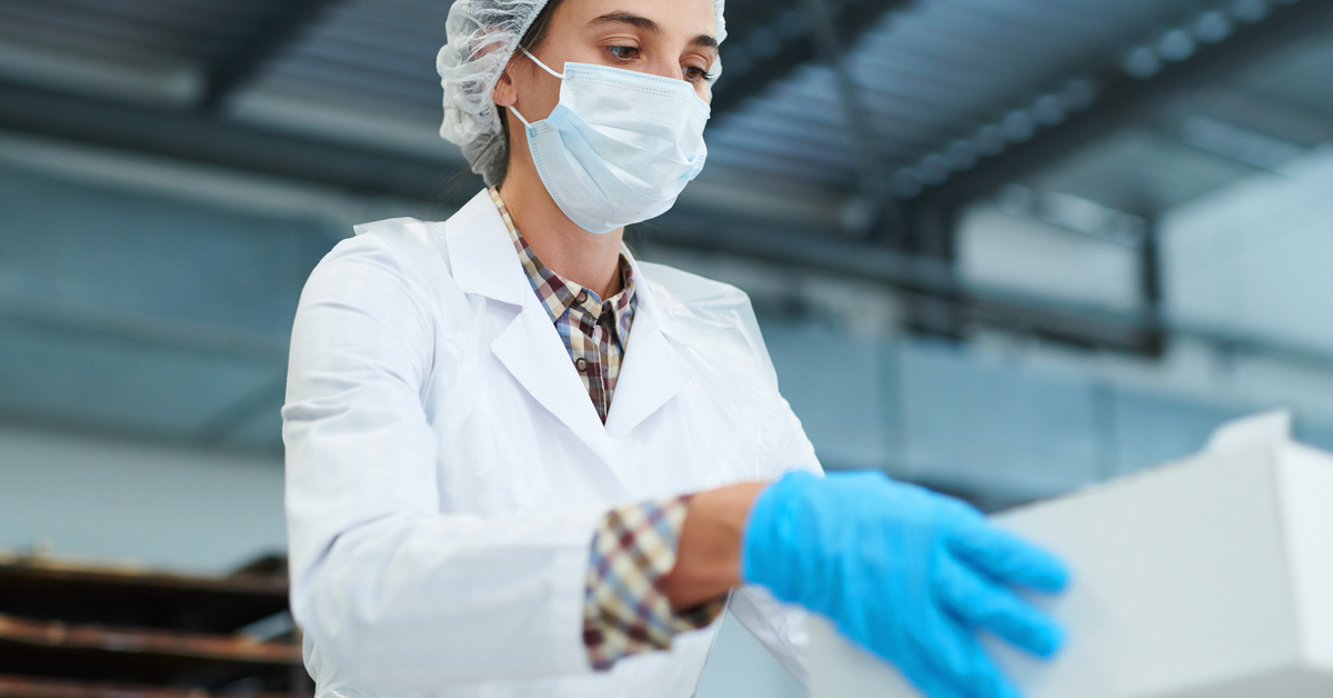An employee wearing a mask and blue gloves is handling a white cardboard box. The box has a clear matte sheen.