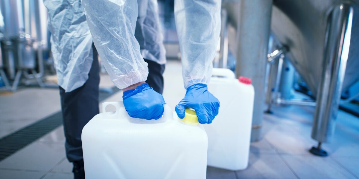 A lab worker wearing appropriate safety gloves is bottling up hazardous chemicals. The large jug is resting firmly on the floor.