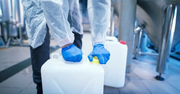 A lab worker wearing appropriate safety gloves is bottling up hazardous chemicals. The large jug is resting firmly on the floor.