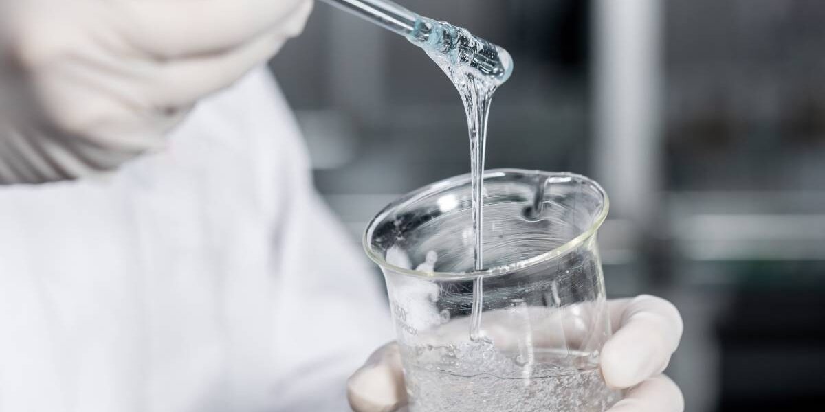 Gloved hands hold a beaker full of a thick, clear liquid. One hand is using a swab to lift some of the liquid out of the glass.