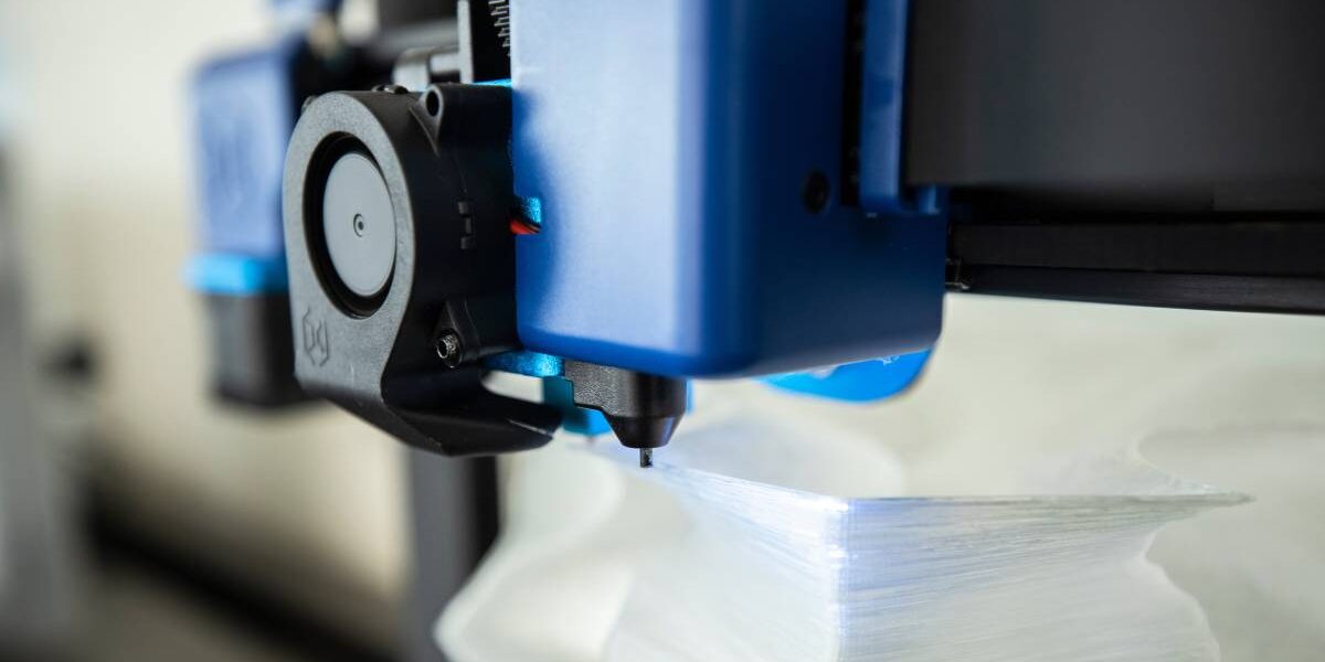 Closeup on the nozzle of a 3D printer. The blue head of the printer uses a black nozzle to print out a white figure.