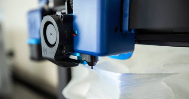 Closeup on the nozzle of a 3D printer. The blue head of the printer uses a black nozzle to print out a white figure.