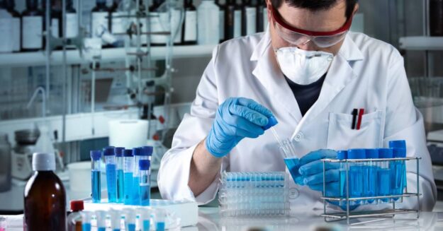 A researcher wearing goggles and a mask works in a lab setting with numerous blue chemicals in test tubes.