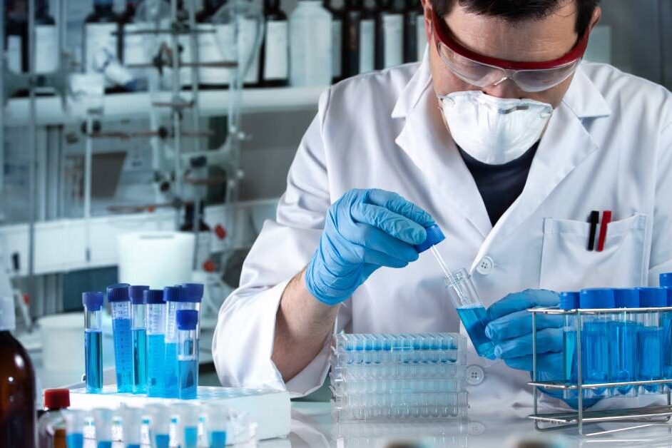 A researcher wearing goggles and a mask works in a lab setting with numerous blue chemicals in test tubes.