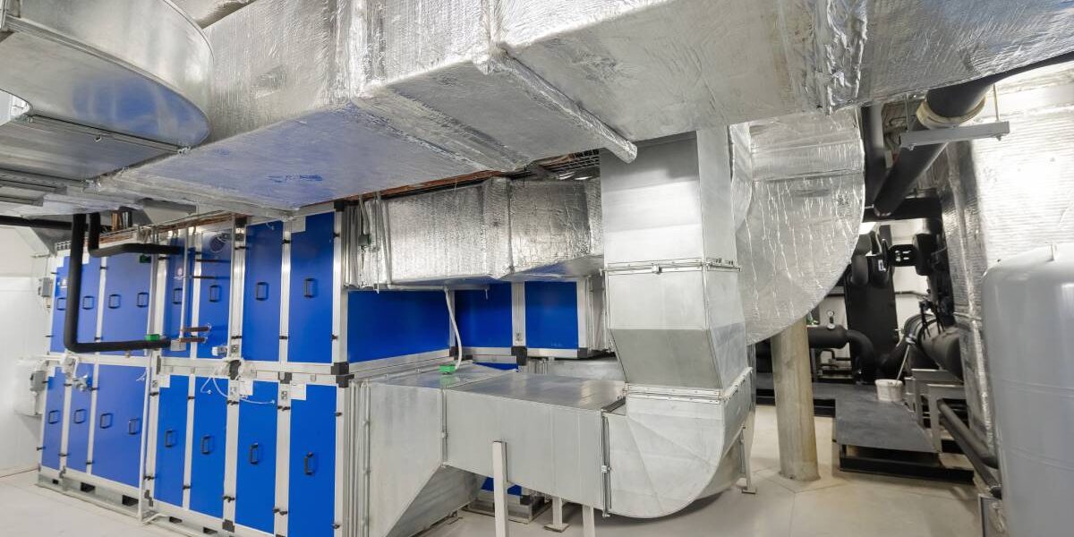 Multiple ducts leading to a structure with multiple blue doors in an indoor industrial facility with pipes.