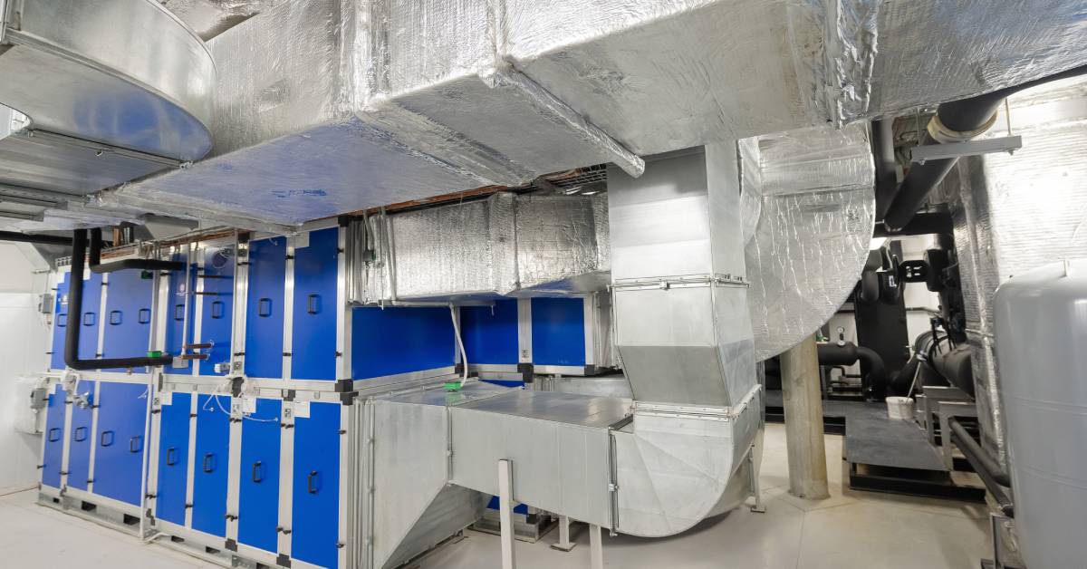 Multiple ducts leading to a structure with multiple blue doors in an indoor industrial facility with pipes.