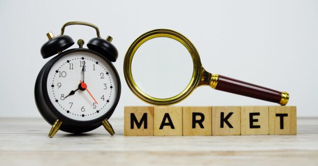 Wooden tiles lined up with letters that spell MARKET with a magnifying glass placed on top and an alarm clock nearby.