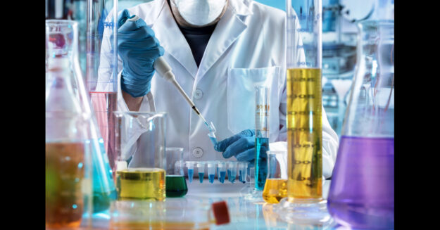 A person wearing a lab coat, face mask, and latex gloves using tools and handling various chemicals in a lab setting.