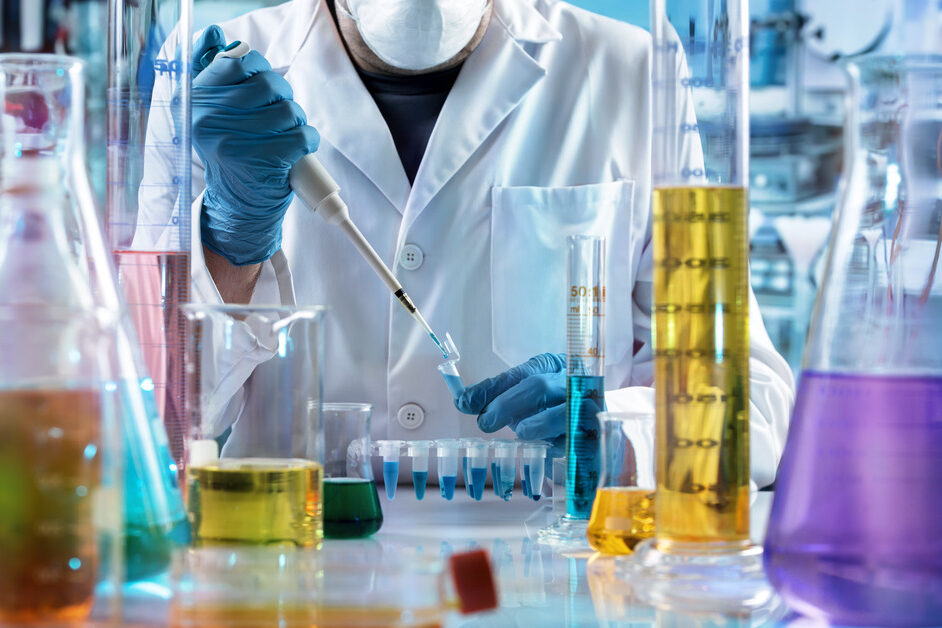A person wearing a lab coat, face mask, and latex gloves using tools and handling various chemicals in a lab setting.