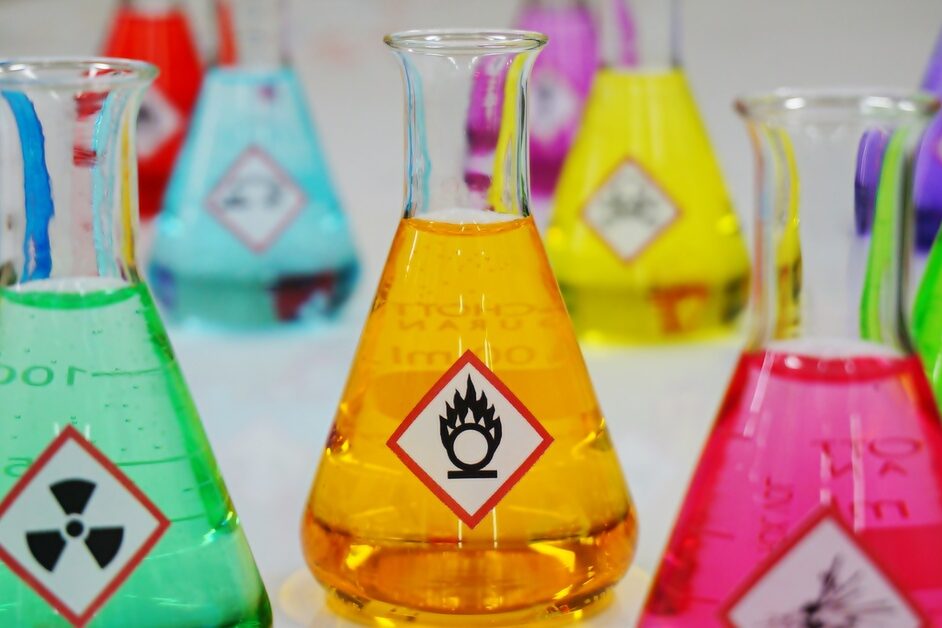 Beakers filled with different-colored liquids and labeled with icons for chemical hazards sit on a white table.