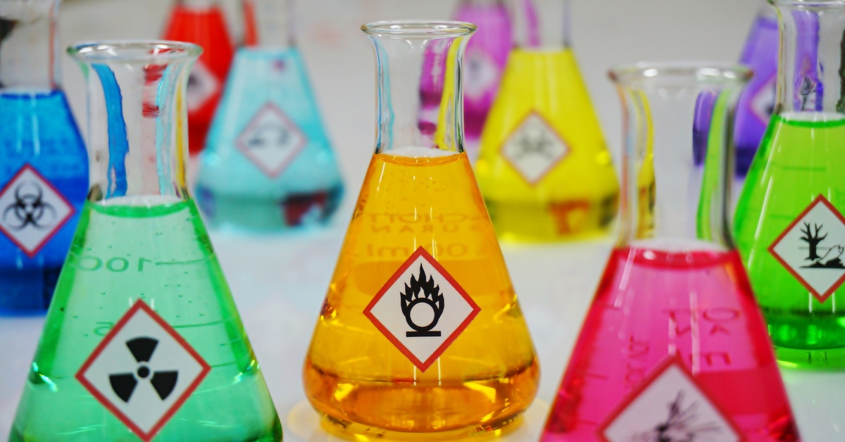 Beakers filled with different-colored liquids and labeled with icons for chemical hazards sit on a white table.