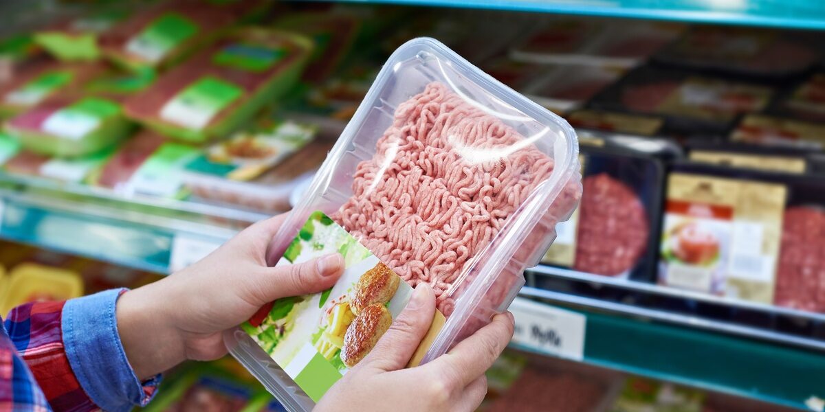A person wearing a long-sleeve flannel shirt holds a plastic package of ground meat in a grocery store. A person wearing a long-sleeve flannel shirt holds a plastic package of ground meat in a grocery store.