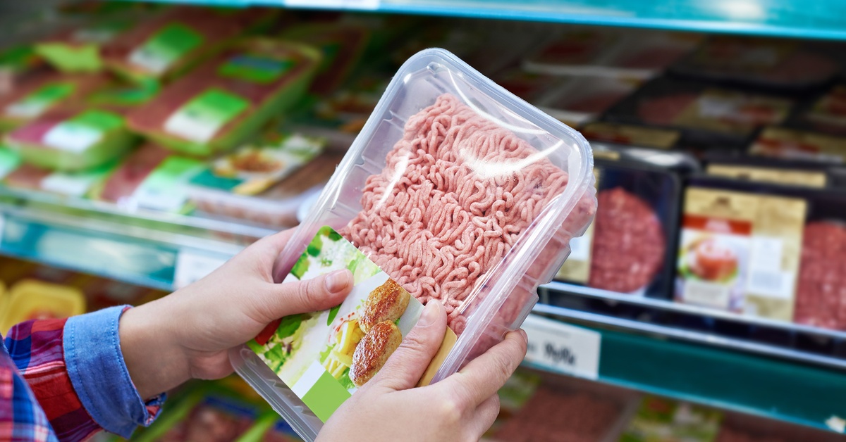A person wearing a long-sleeve flannel shirt holds a plastic package of ground meat in a grocery store.