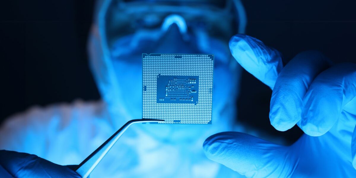 A close-up of a person wearing expansive coveralls, a mask, and goggles using forceps to hold a microprocessor.