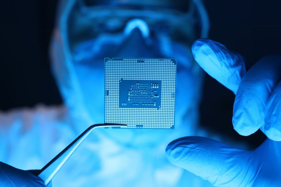 A close-up of a person wearing expansive coveralls, a mask, and goggles using forceps to hold a microprocessor.