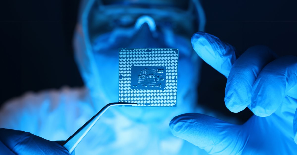 A close-up of a person wearing expansive coveralls, a mask, and goggles using forceps to hold a microprocessor.