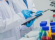 Two people wearing lab coats blue latex gloves, and holding clipboards and pens focus on the information on their papers.