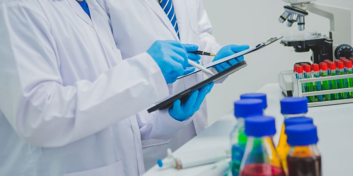 capitalresincorporation-369369-two-lab-techs-Image1 Two people wearing lab coats blue latex gloves, and holding clipboards and pens focus on the information on their papers.