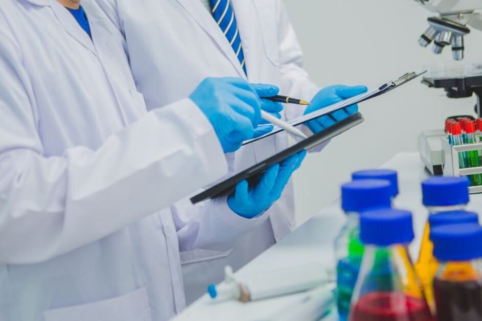 Two people wearing lab coats blue latex gloves, and holding clipboards and pens focus on the information on their papers.