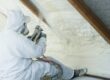A person wearing a white work suit sits down and uses an industrial sprayer to apply polyurethane foam to a roof.