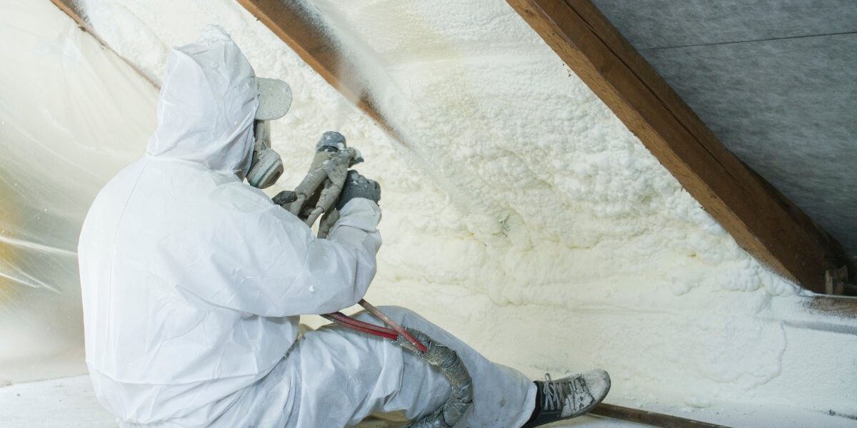 A person wearing a white work suit sits down and uses an industrial sprayer to apply polyurethane foam to a roof.