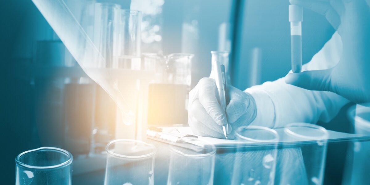 A person wearing gloves holds a test tube in one hand and uses the other to write. The image is washed in blue. A person wearing gloves holds a test tube in one hand and uses the other to write. The image is washed in blue.