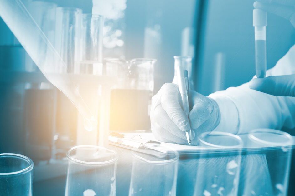 A person wearing gloves holds a test tube in one hand and uses the other to write. The image is washed in blue.