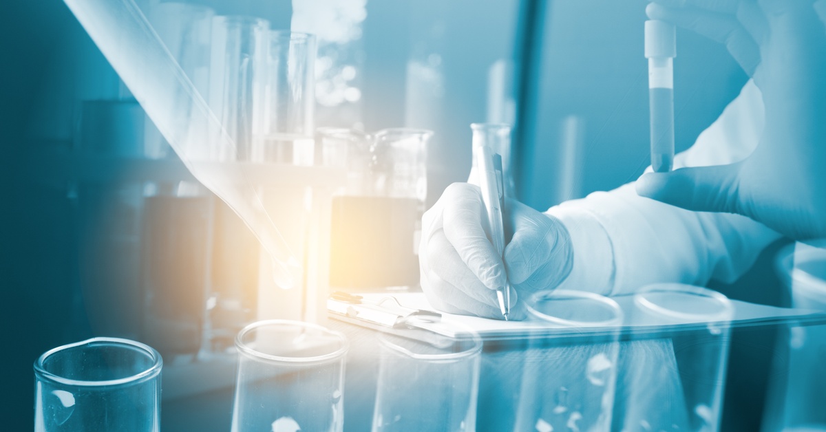 A person wearing gloves holds a test tube in one hand and uses the other to write. The image is washed in blue.