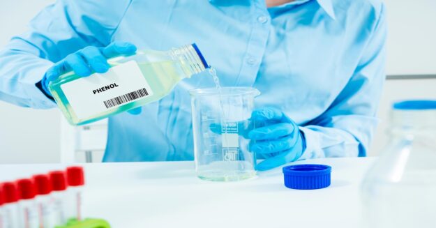 A person wearing blue gloves pours liquid from a clear container labeled "phenol" into another clear container.