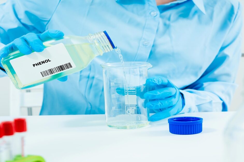 A person wearing blue gloves pours liquid from a clear container labeled "phenol" into another clear container.