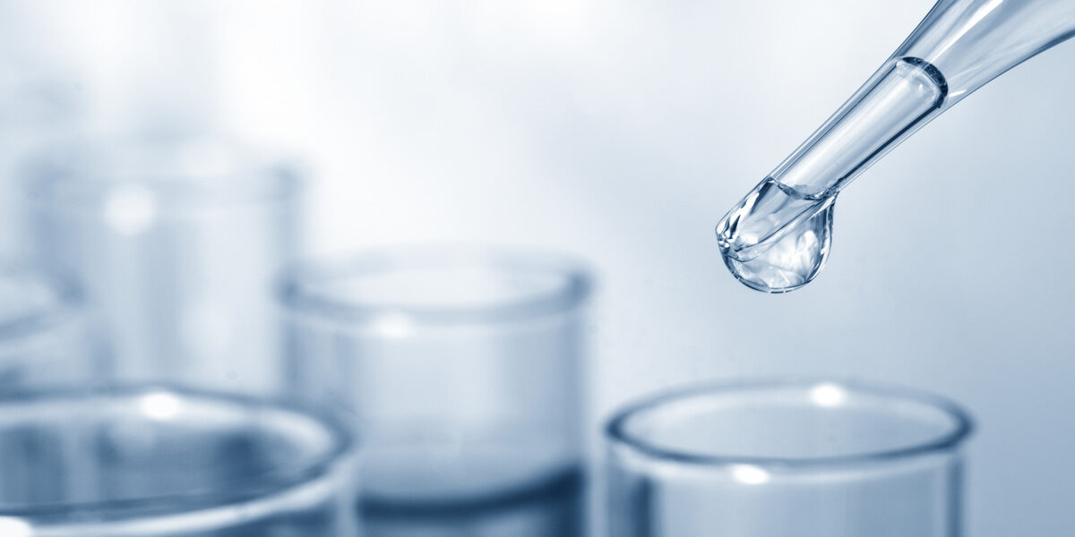 A drop of clear liquid forming at the tip of a pipette and hanging over the top of a glass tube placed among other glassware.