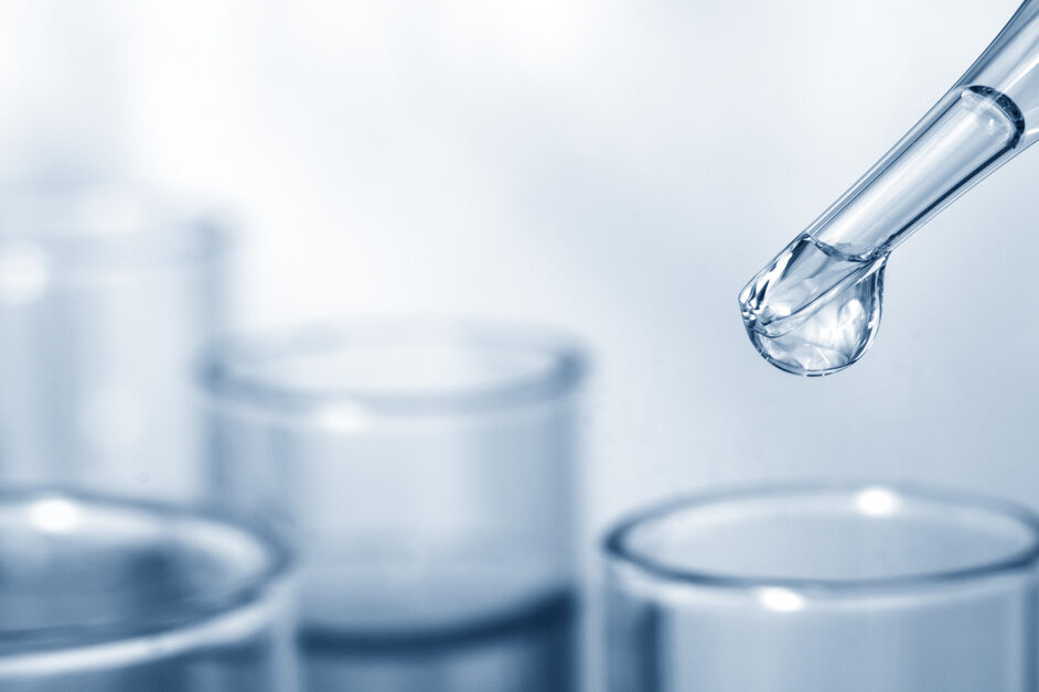 A drop of clear liquid forming at the tip of a pipette and hanging over the top of a glass tube placed among other glassware.