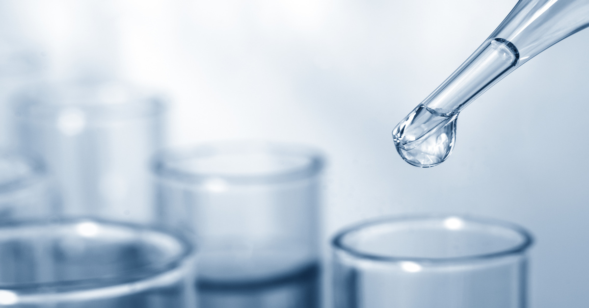 A drop of clear liquid forming at the tip of a pipette and hanging over the top of a glass tube placed among other glassware.