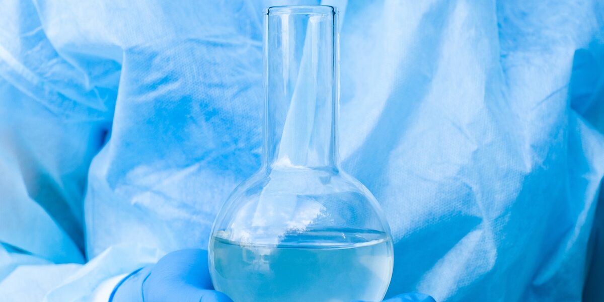 A person wearing a blue glove holds a boiling flask in the palm of their hand. The flask contains liquid. A person wearing a blue glove holds a boiling flask in the palm of their hand. The flask contains liquid.