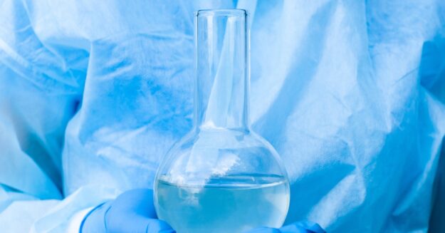 A person wearing a blue glove holds a boiling flask in the palm of their hand. The flask contains liquid.