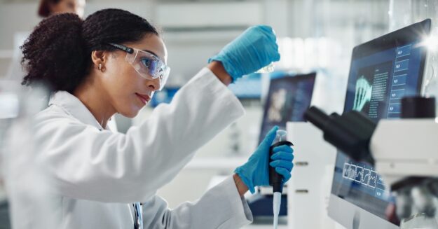 A scientist wearing safety goggles and blue latex gloves is using a pipette in front of a microscope and computer monitor.
