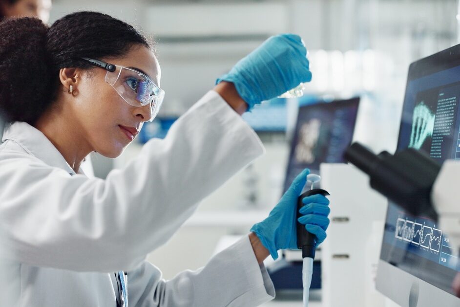 A scientist wearing safety goggles and blue latex gloves is using a pipette in front of a microscope and computer monitor.