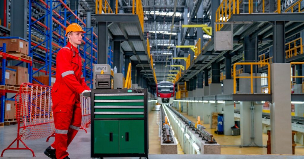 A person wearing red coveralls and a yellow hard hat pushes a tool cart with caster wheels in a large industrial setting.