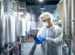 A person wearing a hairnet, face mask, and protective clothing uses a tablet in a chemical manufacturing facility.