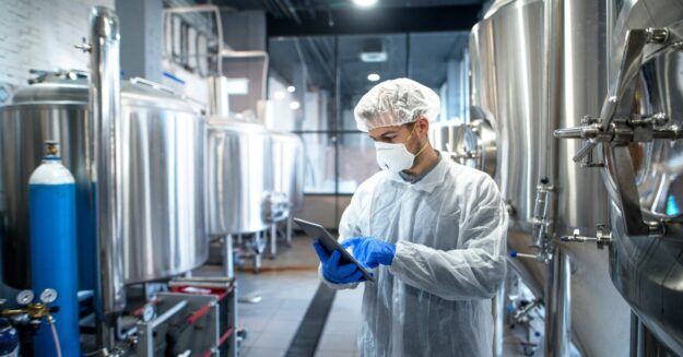 A person wearing a hairnet, face mask, and protective clothing uses a tablet in a chemical manufacturing facility.