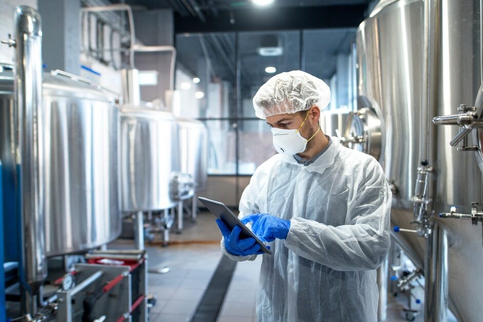 A person wearing a hairnet, face mask, and protective clothing uses a tablet in a chemical manufacturing facility.