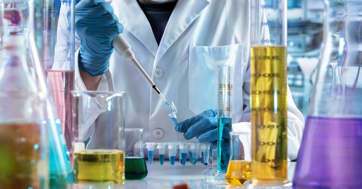 A scientist in a lab coat and gloves uses a micropipette to transfer blue liquid into a test tube on a busy lab bench.