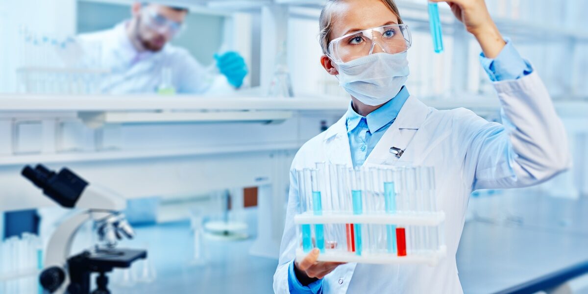 A person wearing safety goggles holds a tray of beakers containing various liquids. They're holding one beaker up to the light.