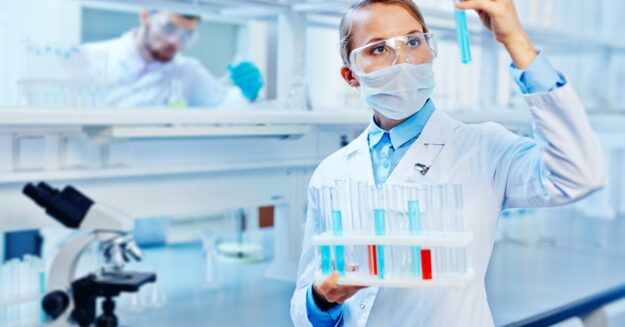 A person wearing safety goggles holds a tray of beakers containing various liquids. They're holding one beaker up to the light.