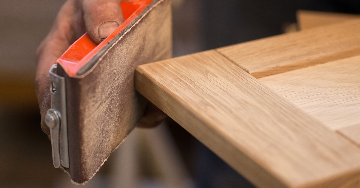 A close-up of a person sanding the corner of a light-colored piece of wood furniture with a sanding block.