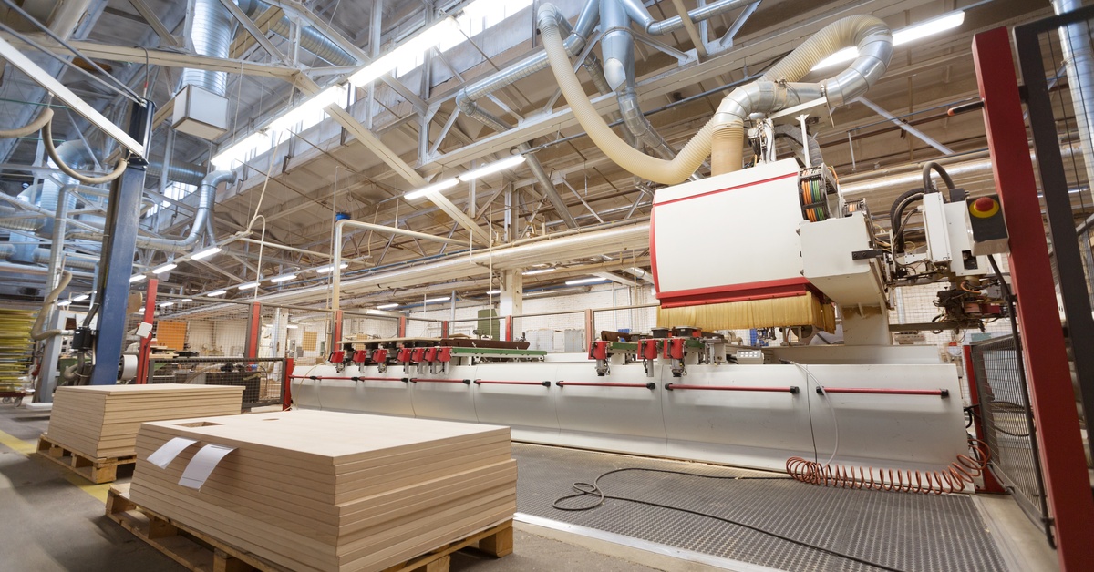 A large woodworking factory with a CNC machine, stacked wooden panels, and an overhead ventilation system.