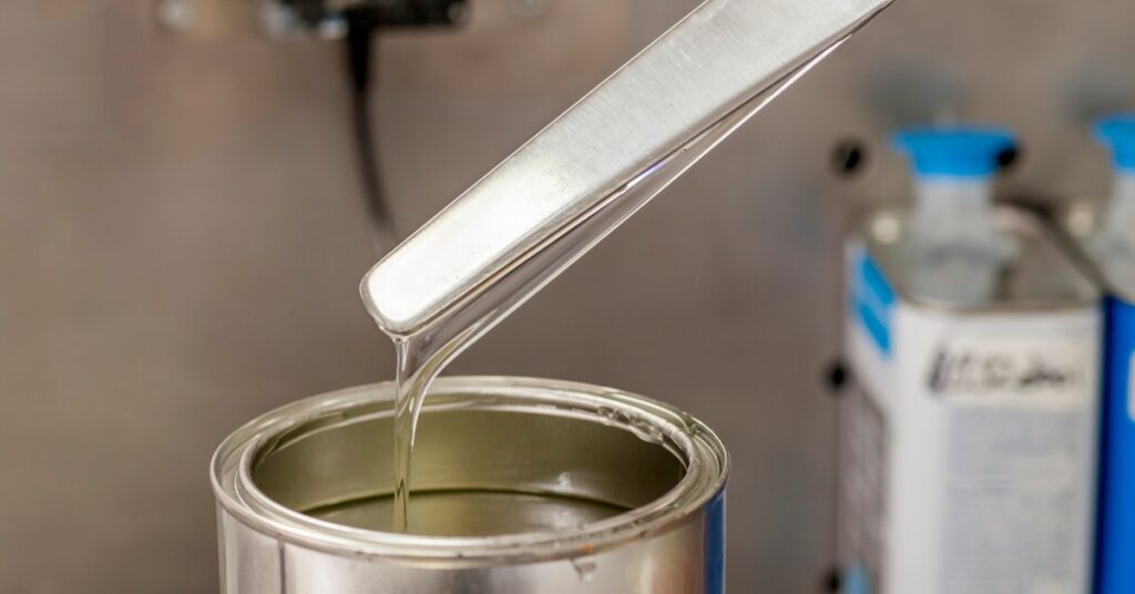A metal spatula hovers above a container of clear resin. A trail of resin drips off the spatula into the bucket.