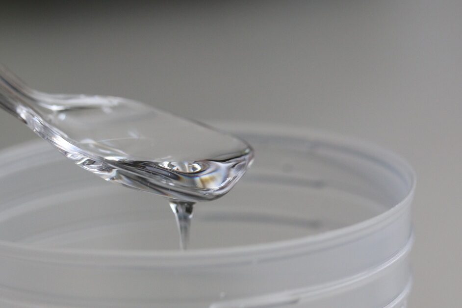 A clear, viscous resin drips from a transparent spoon into a shallow plastic container, set against a soft grey background.